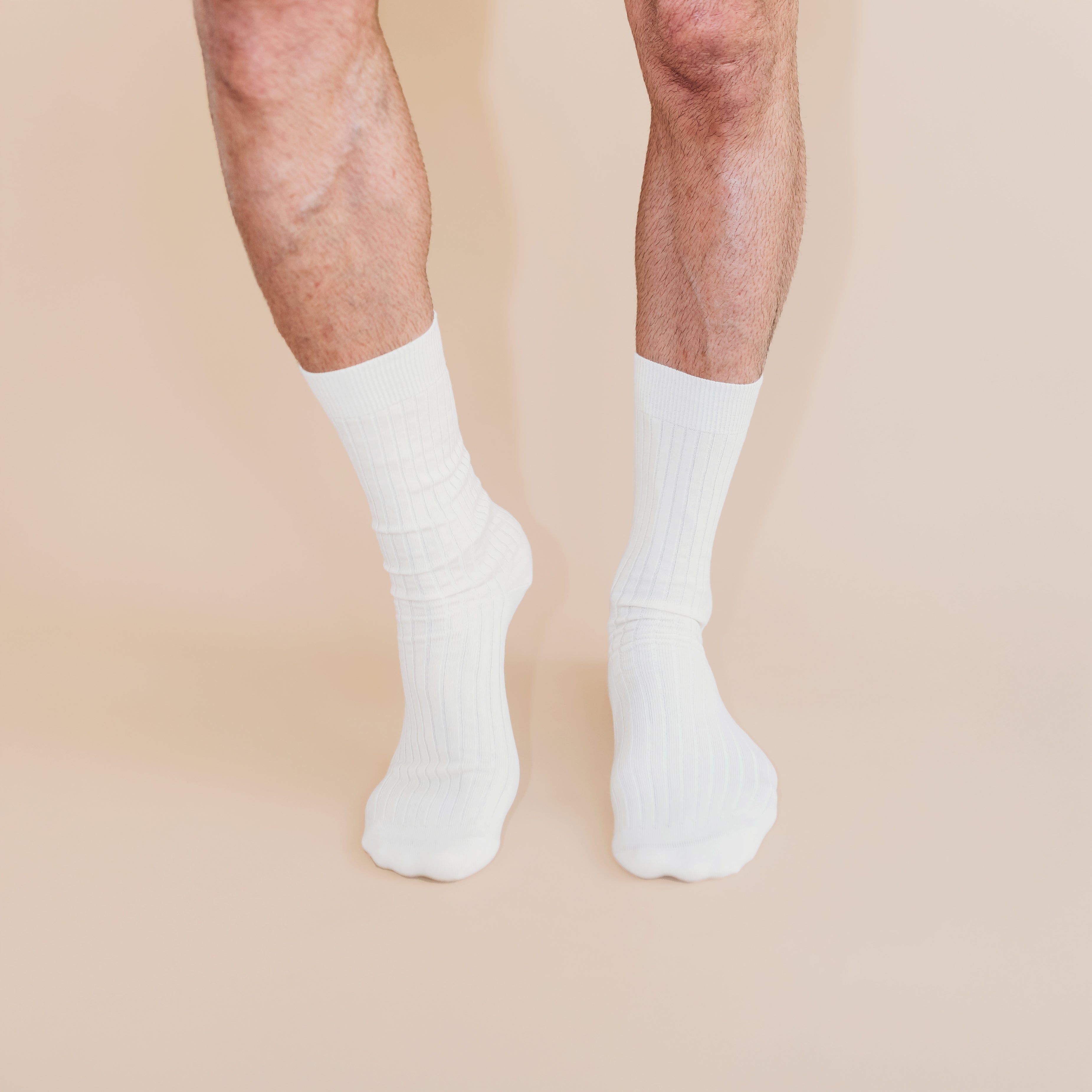 A pair of white ribbed socks on adult legs against a beige background.