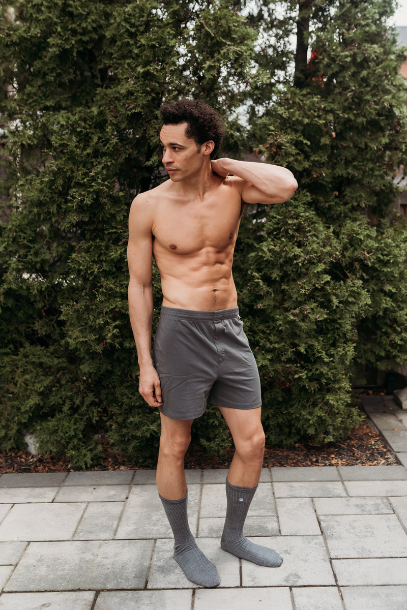 Man wearing gray underwear and socks standing outdoors with greenery in the background