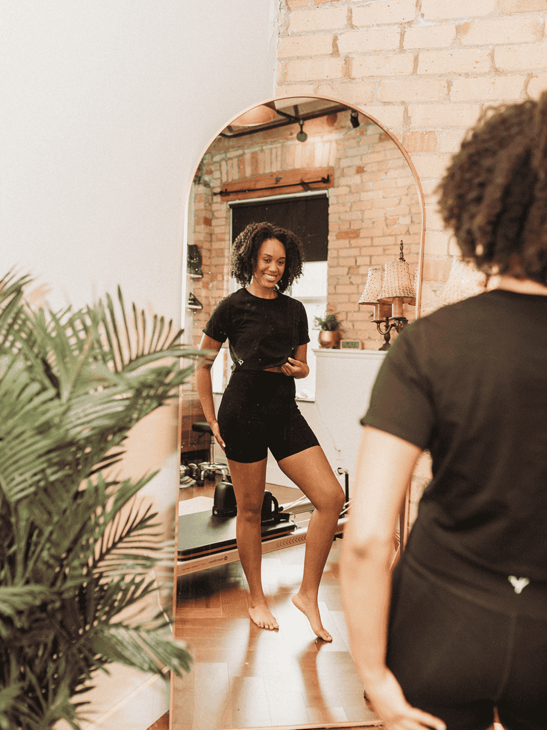 Woman in black outfit standing in front of a mirror in a room with plants and decor.