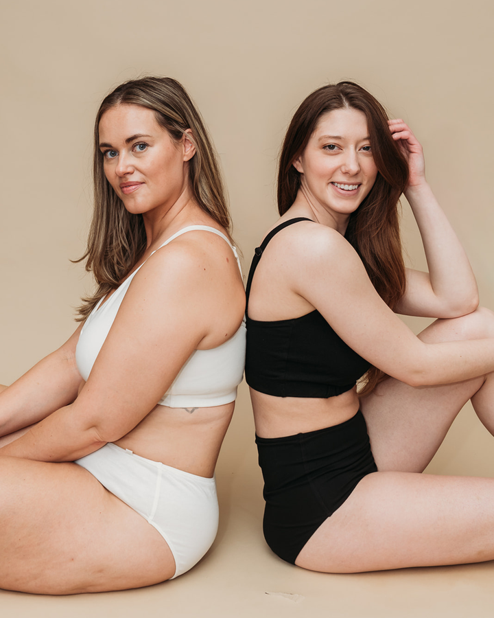 Two women sitting side by side wearing white and black underwear on a beige background
