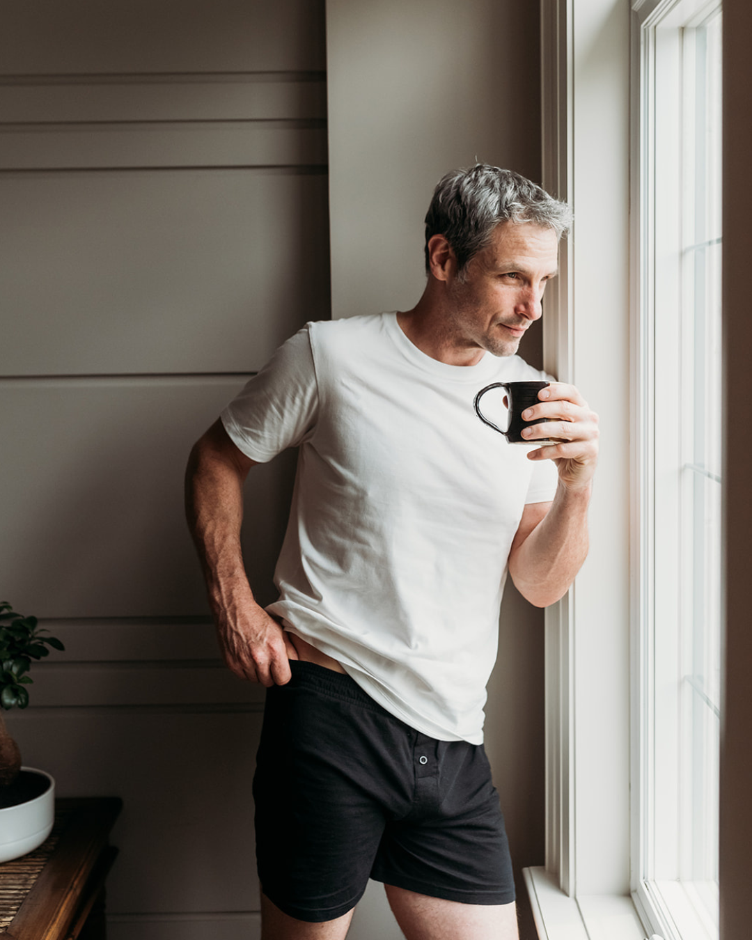 Man in Everyday Knit Organic Cotton Boxers in Black and white tee holding coffee by sunlit window | Q for Quinn