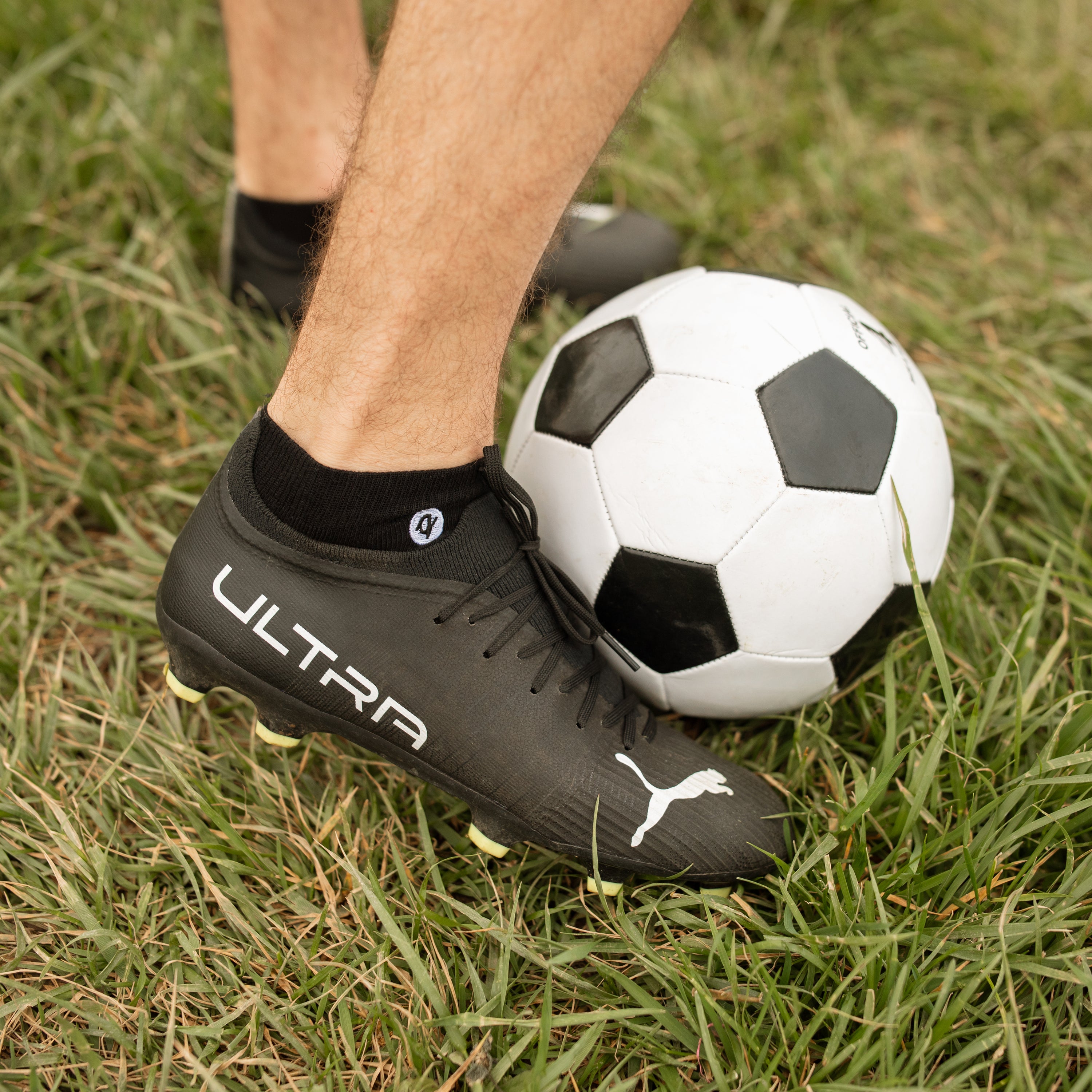Black Organic Cotton Ankle Pacers on feet in soccer cleats beside a football on grass | Q for Quinn