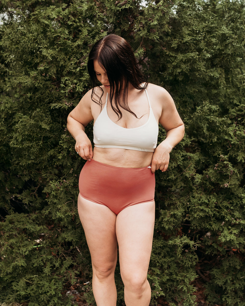 Model wearing Women's High Waist Brief in Dark Madder, outdoors among green evergreen shrubs | Q for Quinn