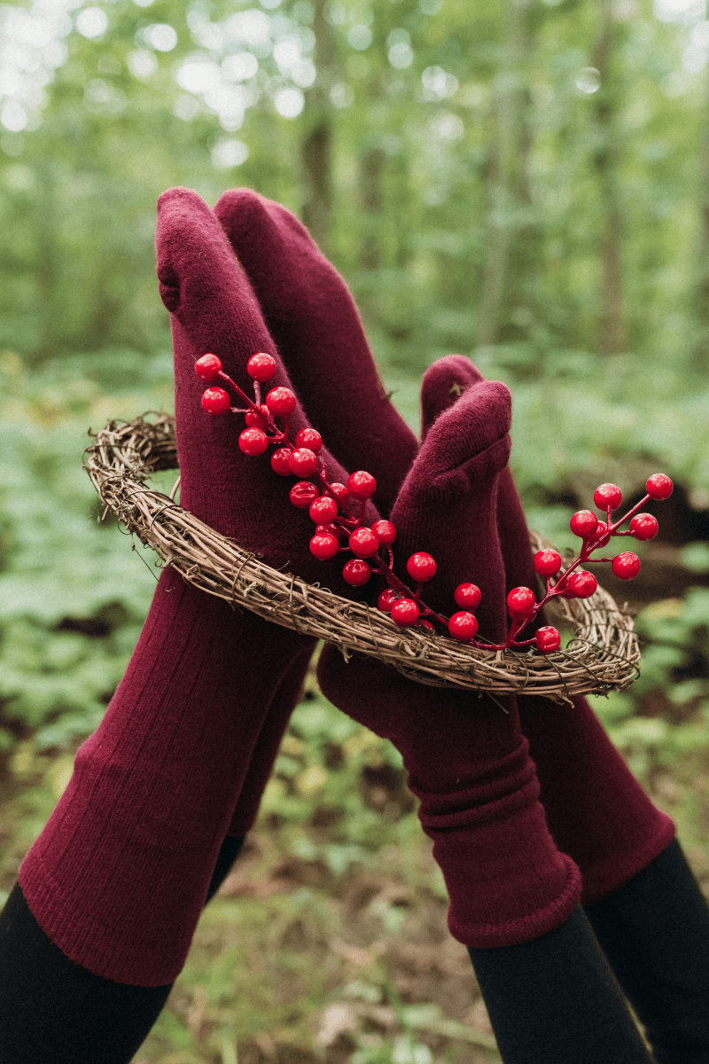 A pair of maroon knit gloves worn on hands holding a small woven wreath with red berries, photographed outdoors in a blurred green forest setting.
