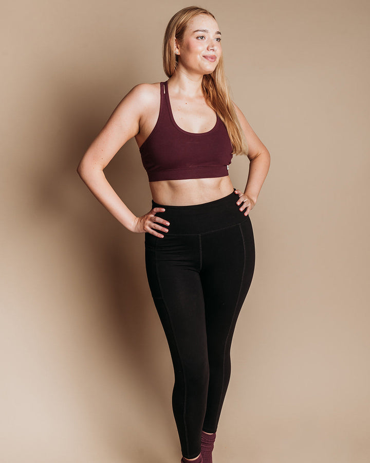 A woman modeling a dark burgundy organic sports bra and black high-waisted leggings against a solid beige studio background.