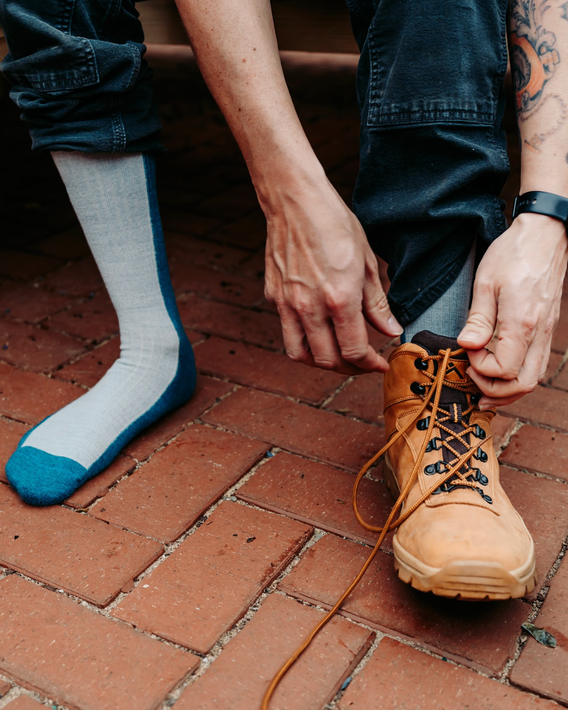 Person tying a brown boot on a brick pavement