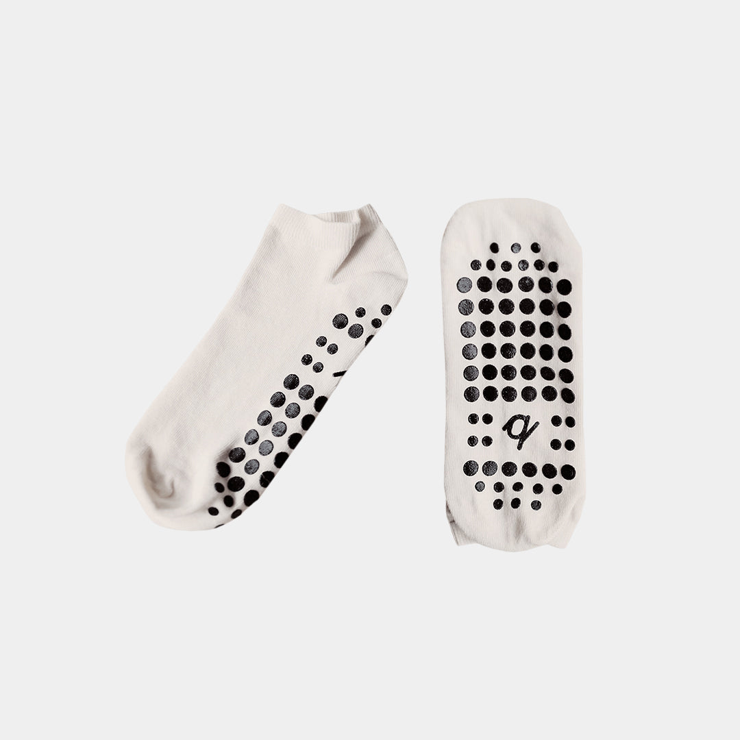 A pair of low-cut white socks with black grip dots on the sole and heel, displayed against a light gray background.