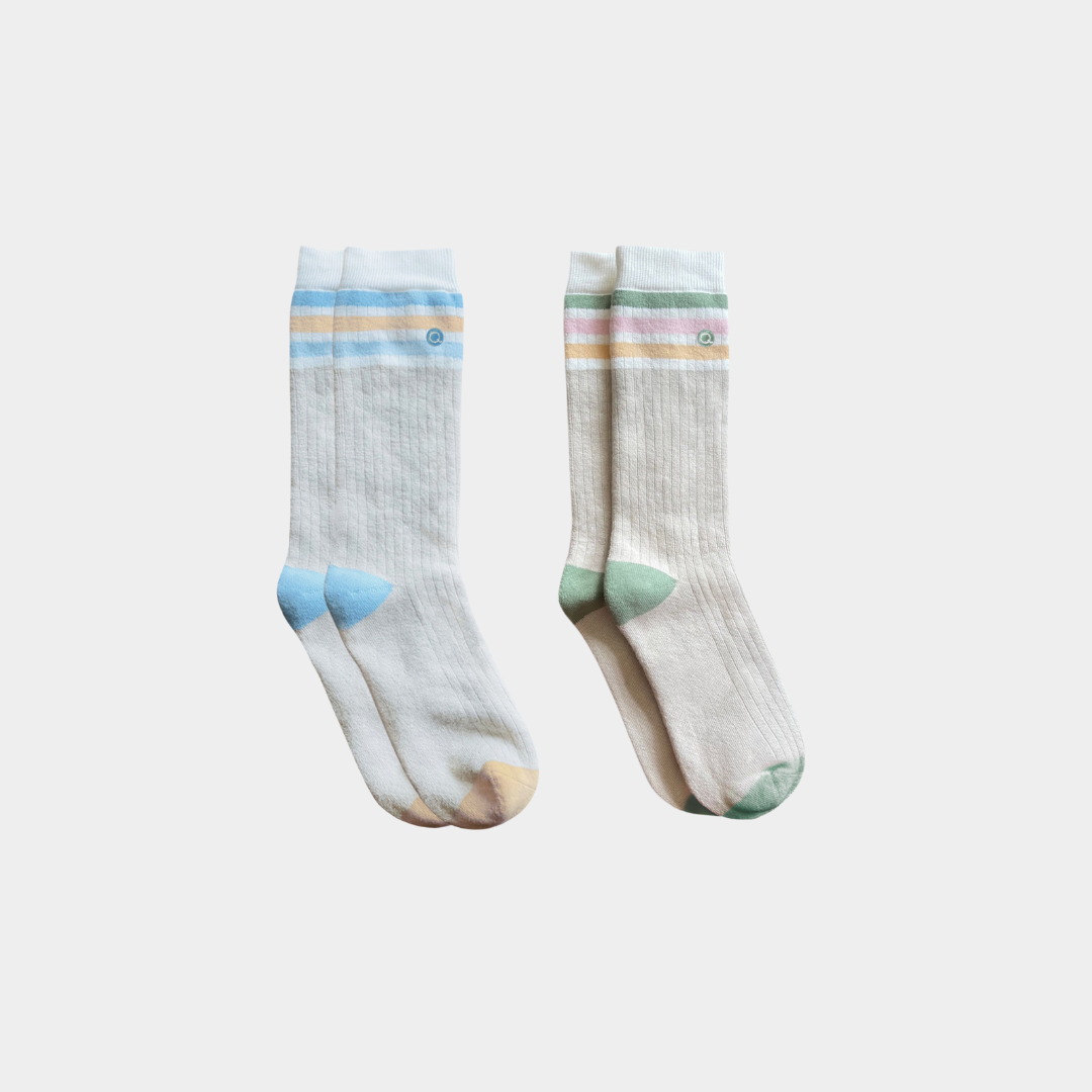 Two pairs of ribbed crew socks in a light cream/off-white color, featuring pastel striped cuffs and multi-colored toe and heel accents in blue, green, and peach, displayed against a light gray background.