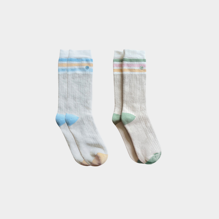 Two pairs of ribbed crew socks in a light cream/off-white color, featuring pastel striped cuffs and multi-colored toe and heel accents in blue, green, and peach, displayed against a light gray background.