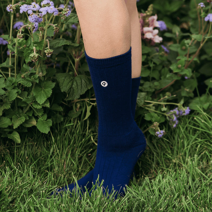A person wearing dark navy blue crew socks standing in green grass with purple flowers in the background. A small circular logo is visible on the side of the front sock.