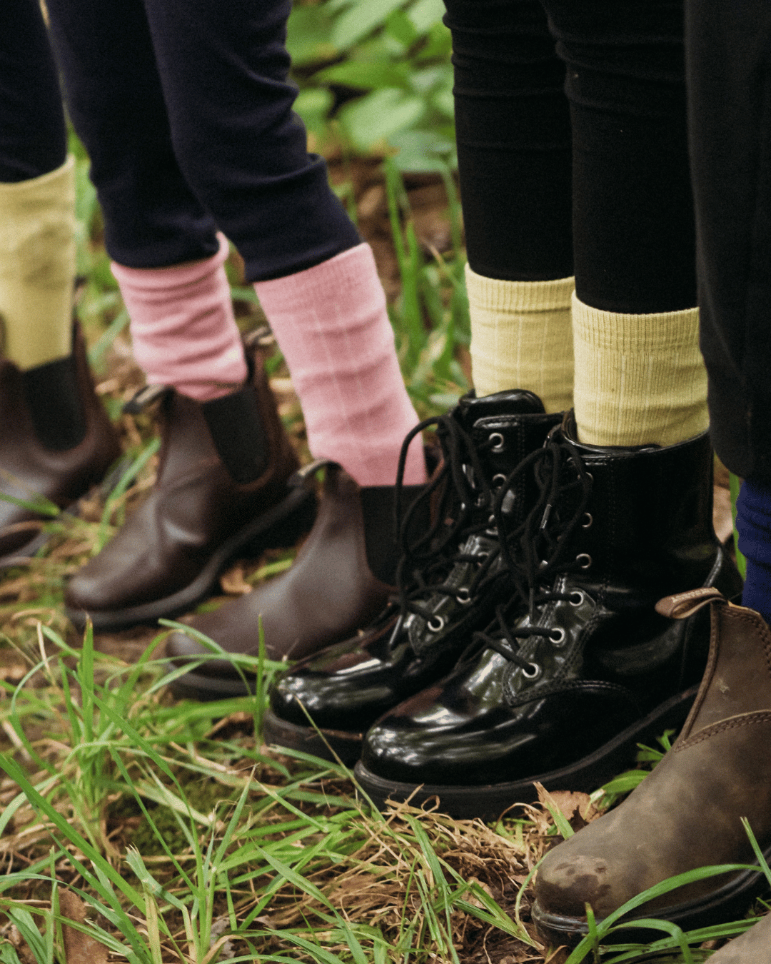 Close-up of several people's lower legs standing outdoors on grass and dirt, wearing different styles of ankle boots and socks. The central focus is on a pair of shiny black lace-up boots with black socks, surrounded by brown boots, pink socks, and pale yellow socks.