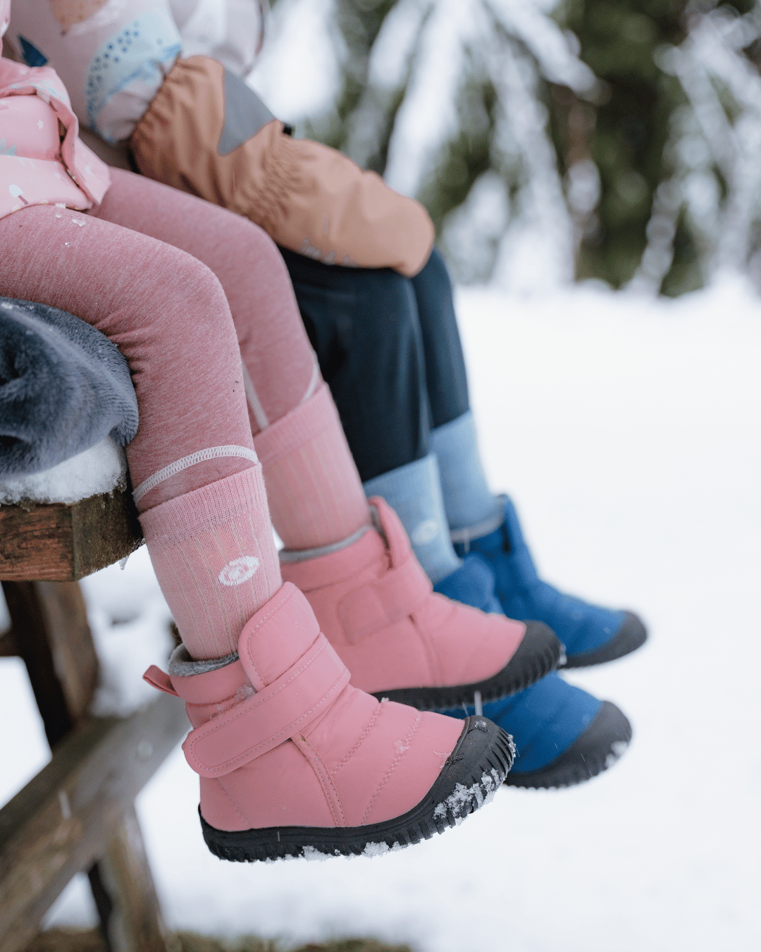 A close-up outdoor winter scene showing children sitting on a bench in the snow, wearing pink and blue winter boots with socks and jackets. The background is a blurred snowy forest.