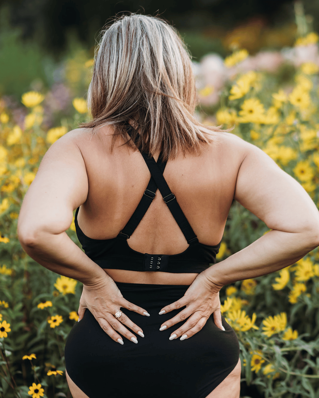 A woman viewed from behind wearing a black sports bra and matching high-waisted leggings, standing in a field of yellow flowers.