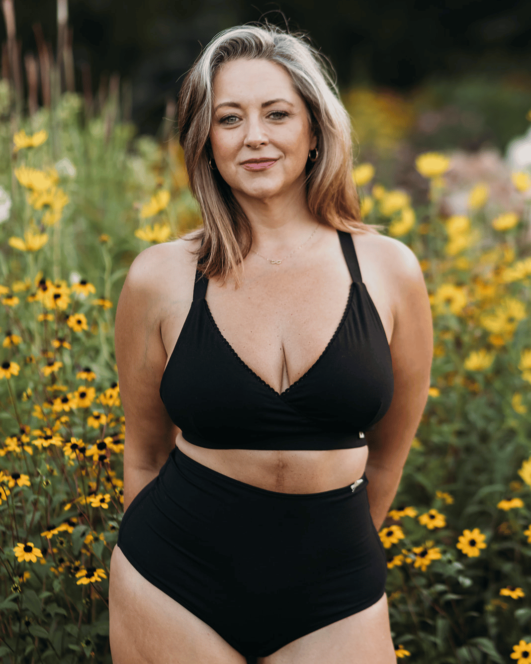 A woman wearing a black two-piece swimsuit stands outdoors in a field of yellow flowers. The set includes a halter-style bikini top with scalloped edges and high-waisted bottoms.