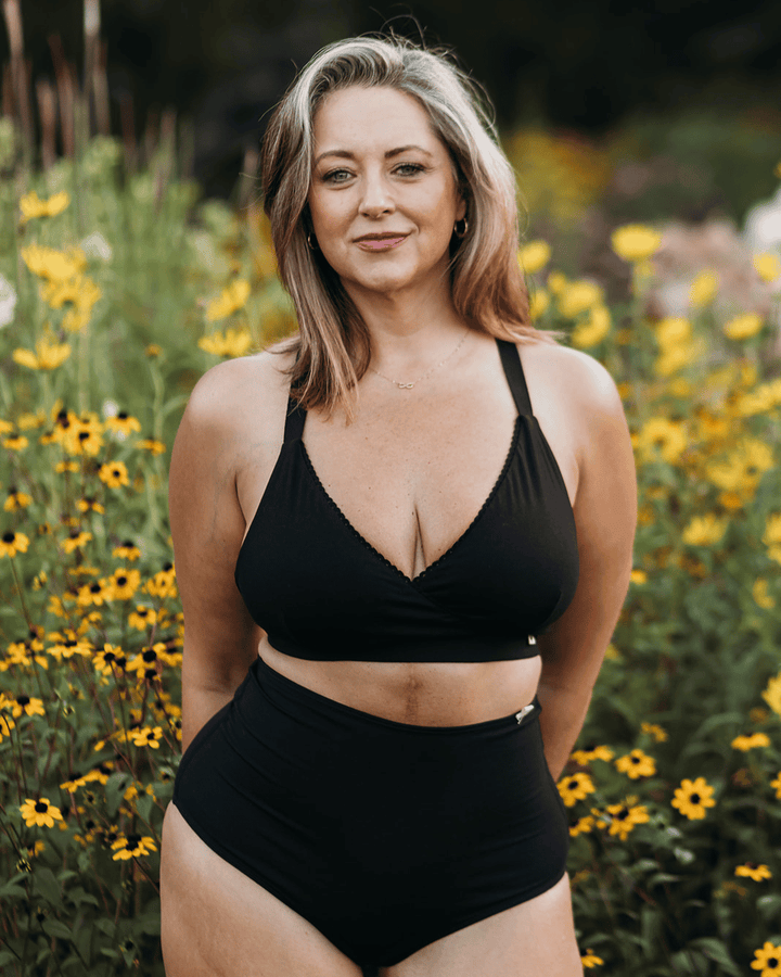 A woman wearing a black two-piece swimsuit stands outdoors in a field of yellow flowers. The set includes a halter-style bikini top with scalloped edges and high-waisted bottoms.