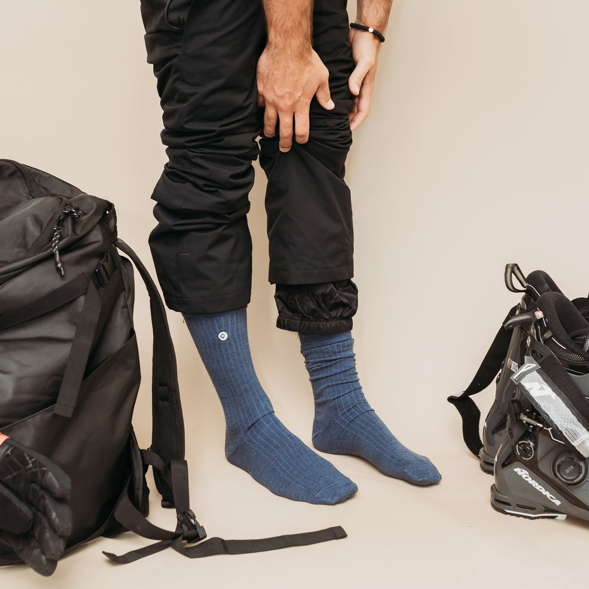 A person wearing black pants and blue ribbed crew socks stands against a plain beige background, with black backpacks and ski gear placed beside them.