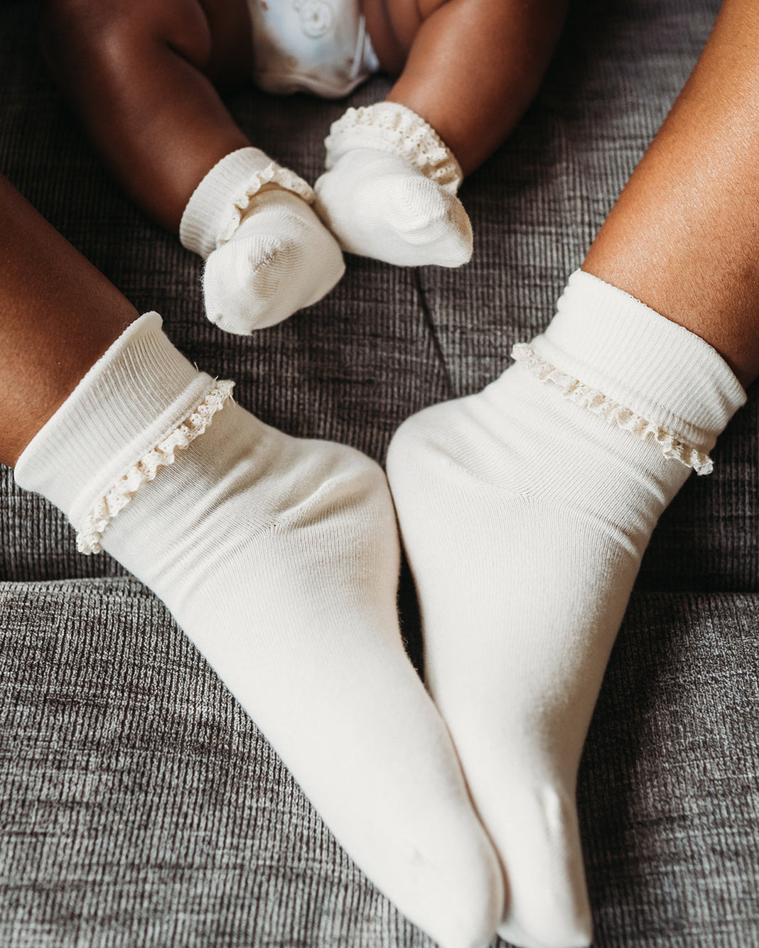 White baby socks with delicate ruffle trim on the cuffs, shown worn on an adult's feet and a baby's feet on a textured gray fabric surface.