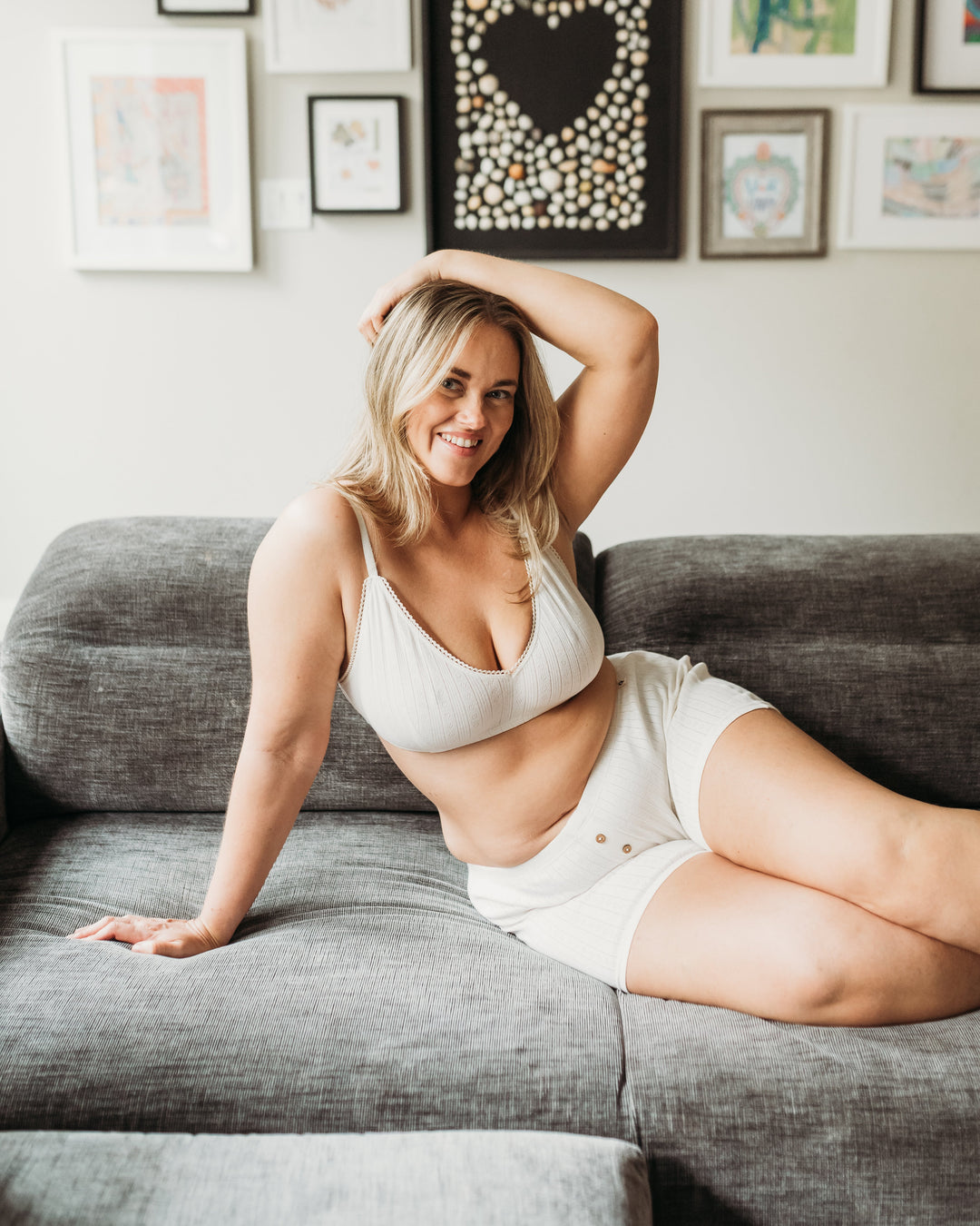 Woman sitting on a gray couch wearing a white bra and shorts, with a gallery wall of framed art in the background.