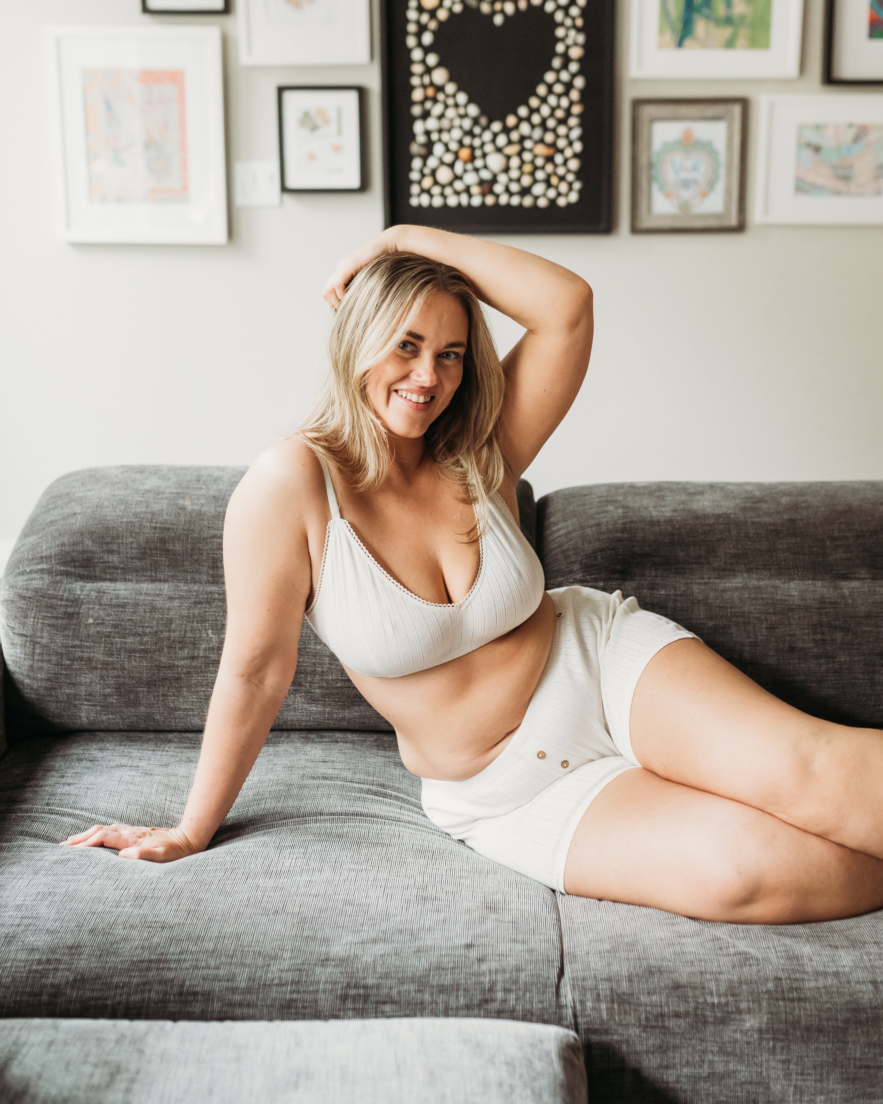 Woman sitting on a gray couch wearing a white bra and shorts, with a gallery wall of framed art in the background.