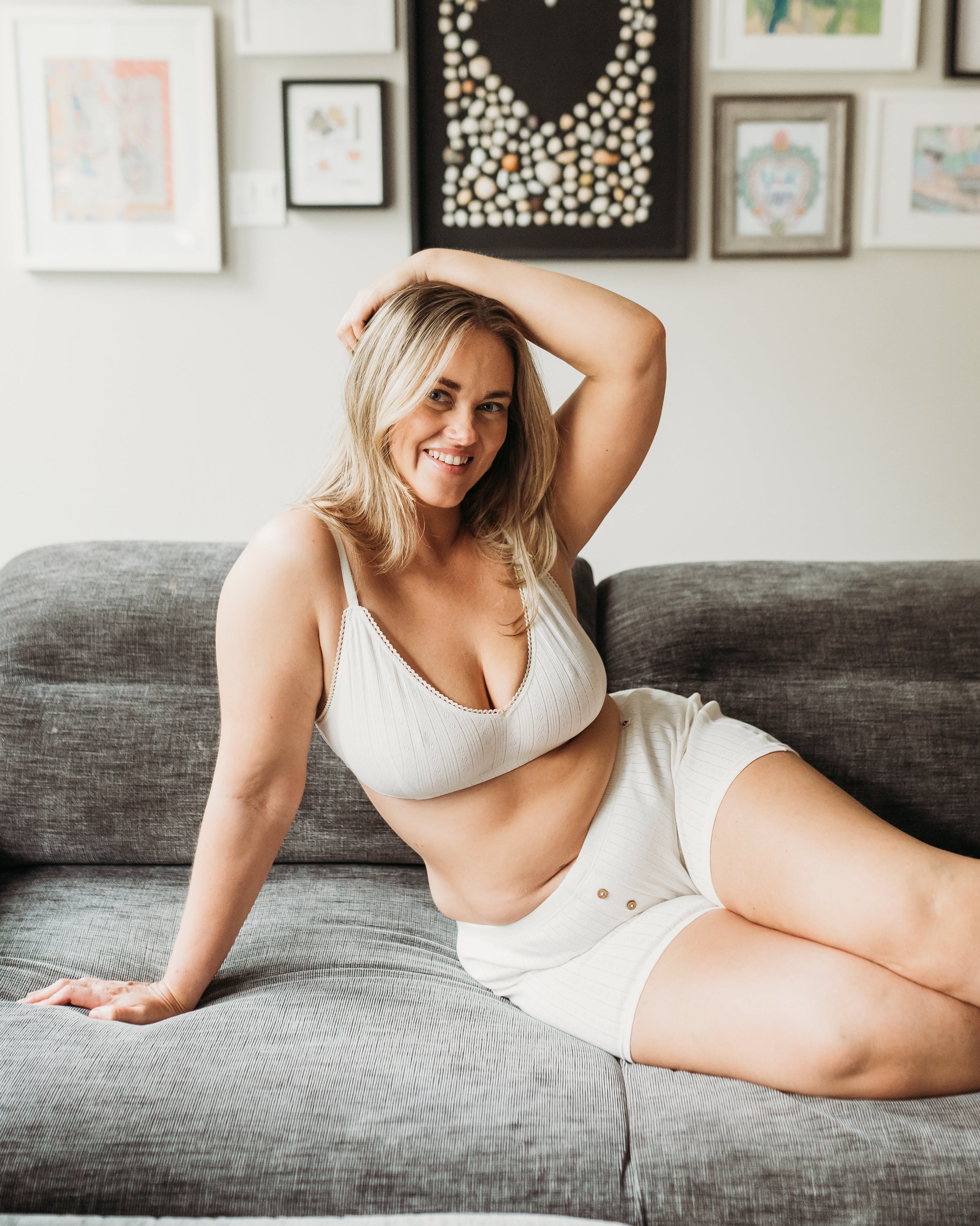 Woman sitting on a gray couch wearing a white bra and shorts, with a gallery wall of framed art in the background.