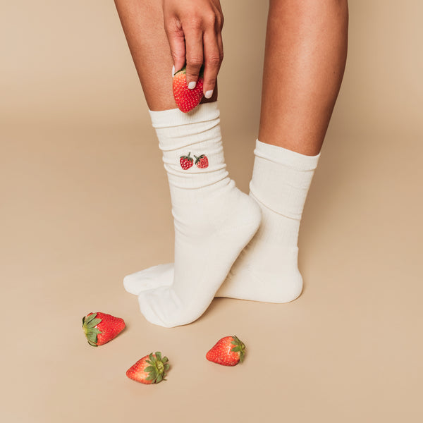 White socks with strawberry design worn by a person, surrounded by strawberries on a beige background
