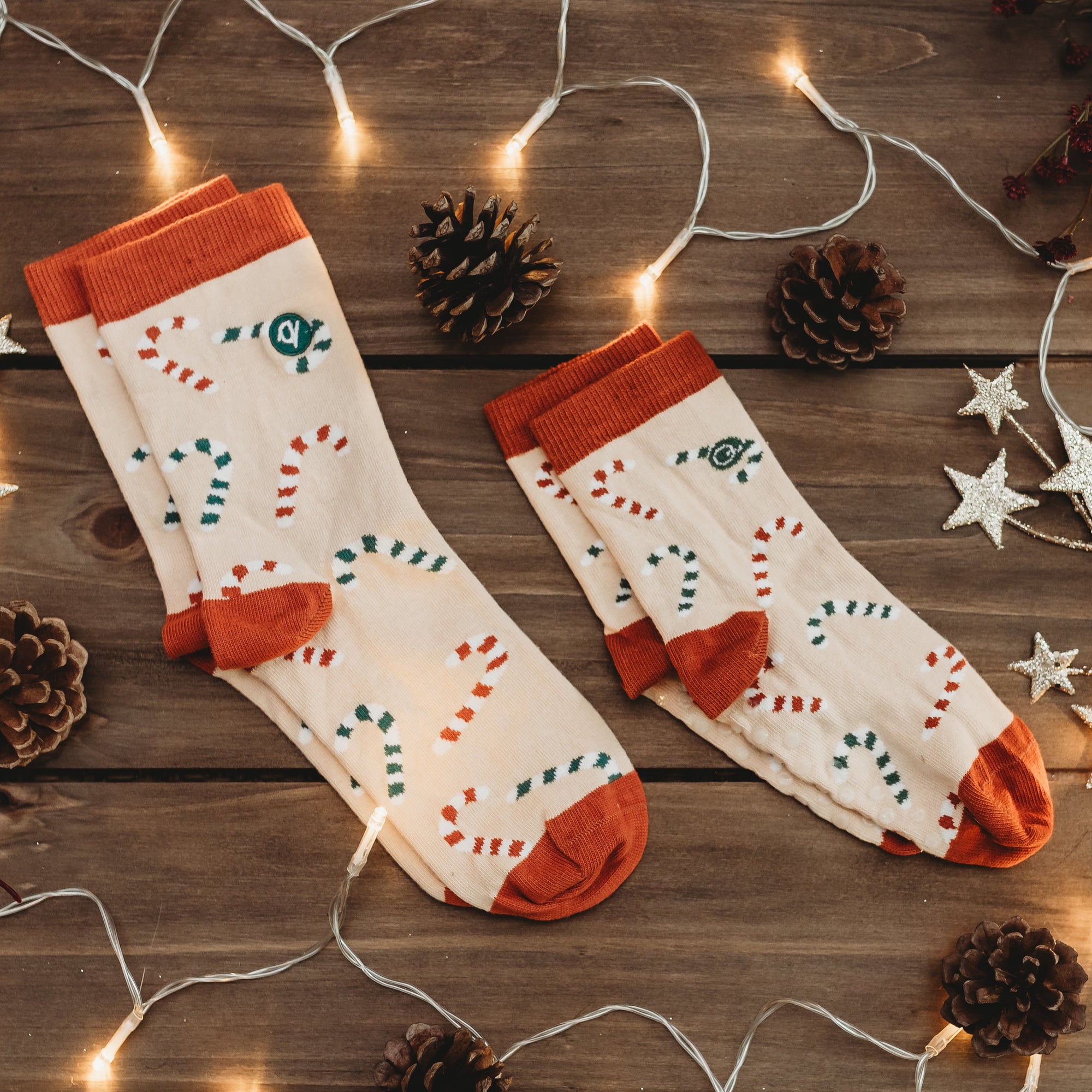 Two socks with orange accents on a wooden surface with decorative lights and pine cones.