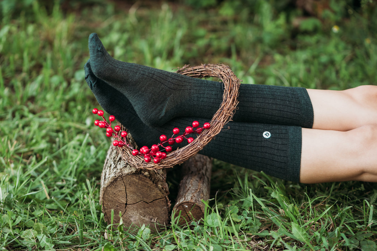 A person lying on grass outdoors wearing dark charcoal ribbed knee-high socks, with one foot resting on a rustic log and a woven wreath decorated with red berries.