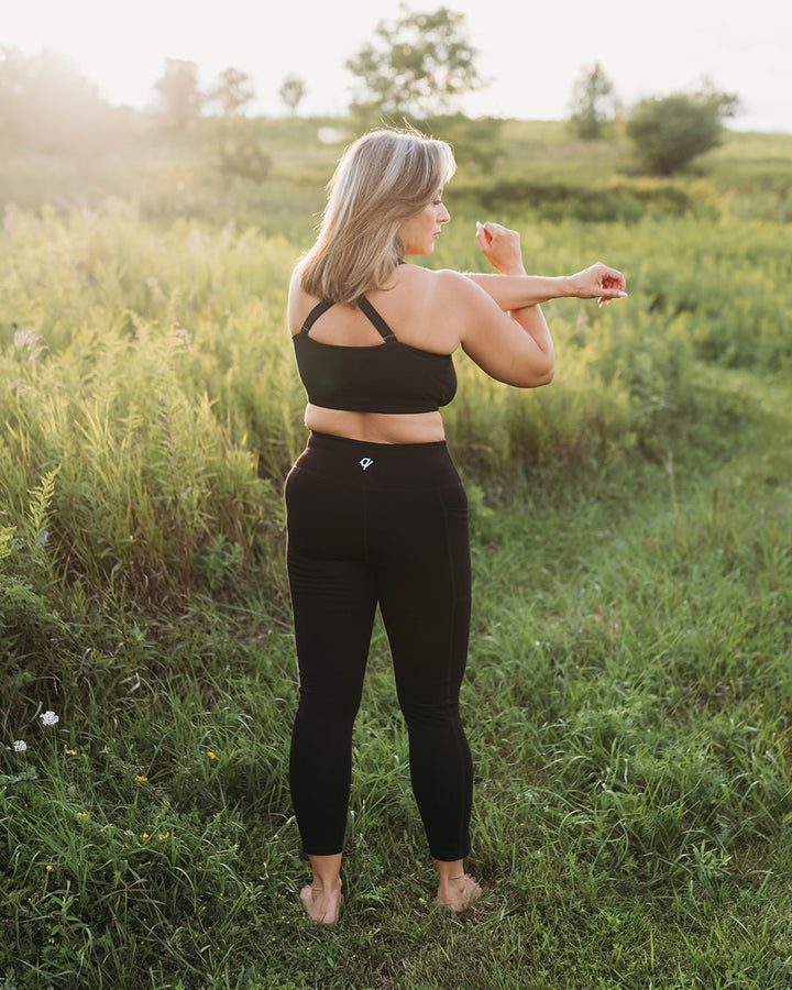 A woman standing in a grassy field at sunset, wearing a black sports bra and black high-waisted leggings, viewed from behind as she stretches her arm.