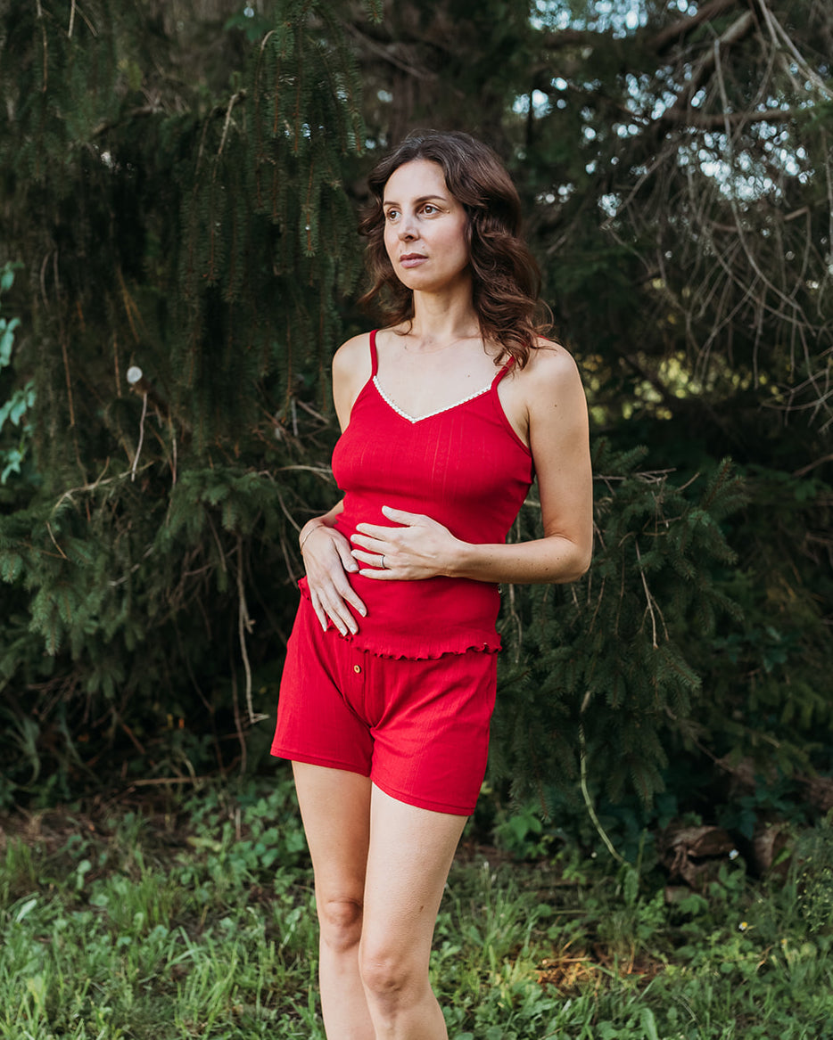 A pregnant woman wearing a matching red two-piece loungewear set consisting of a spaghetti-strap camisole top and shorts, standing outdoors in front of a dense forest background.