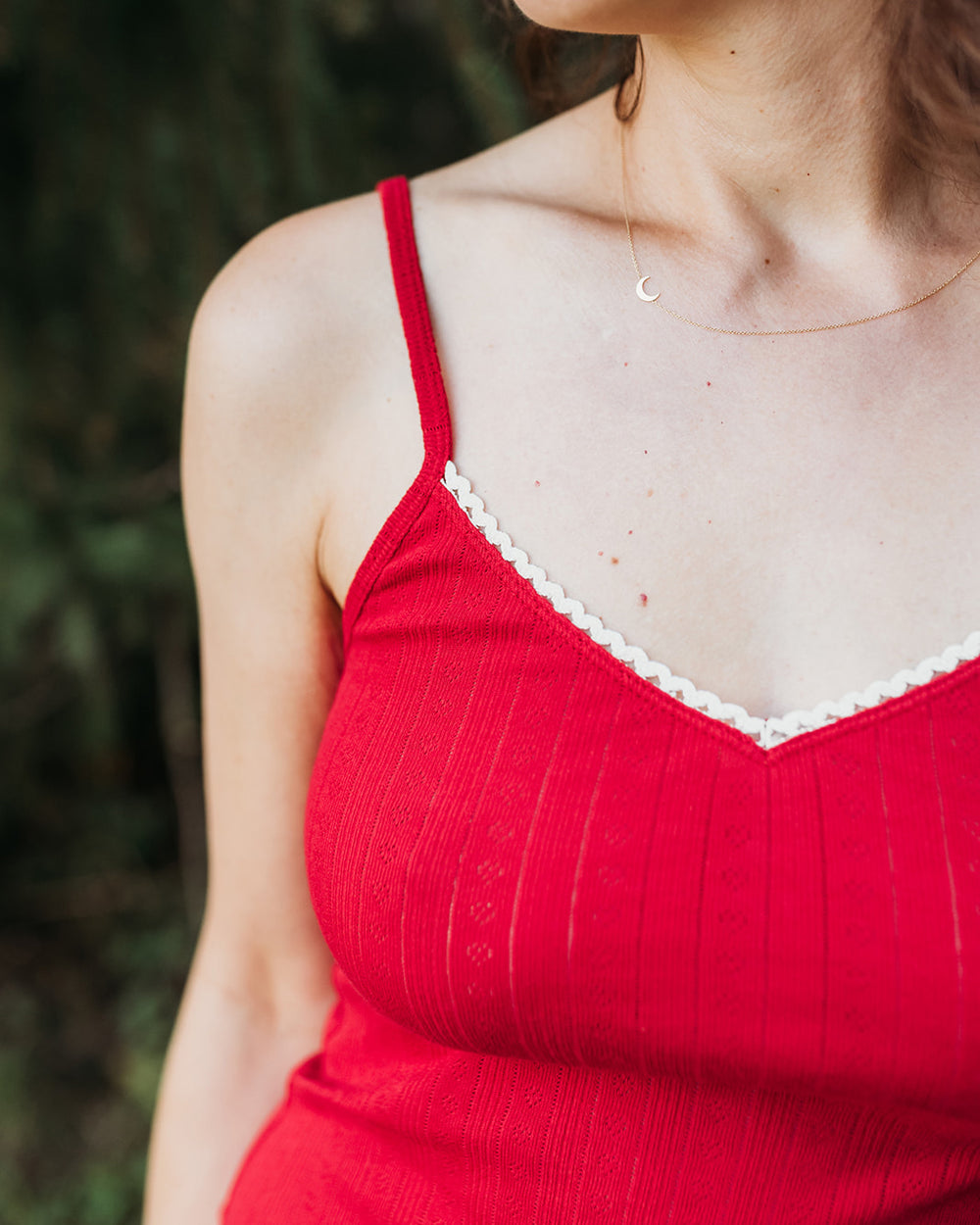 A close-up of a woman wearing a red ribbed camisole top with white scalloped trim along the V-neckline, photographed outdoors against a blurred green background.