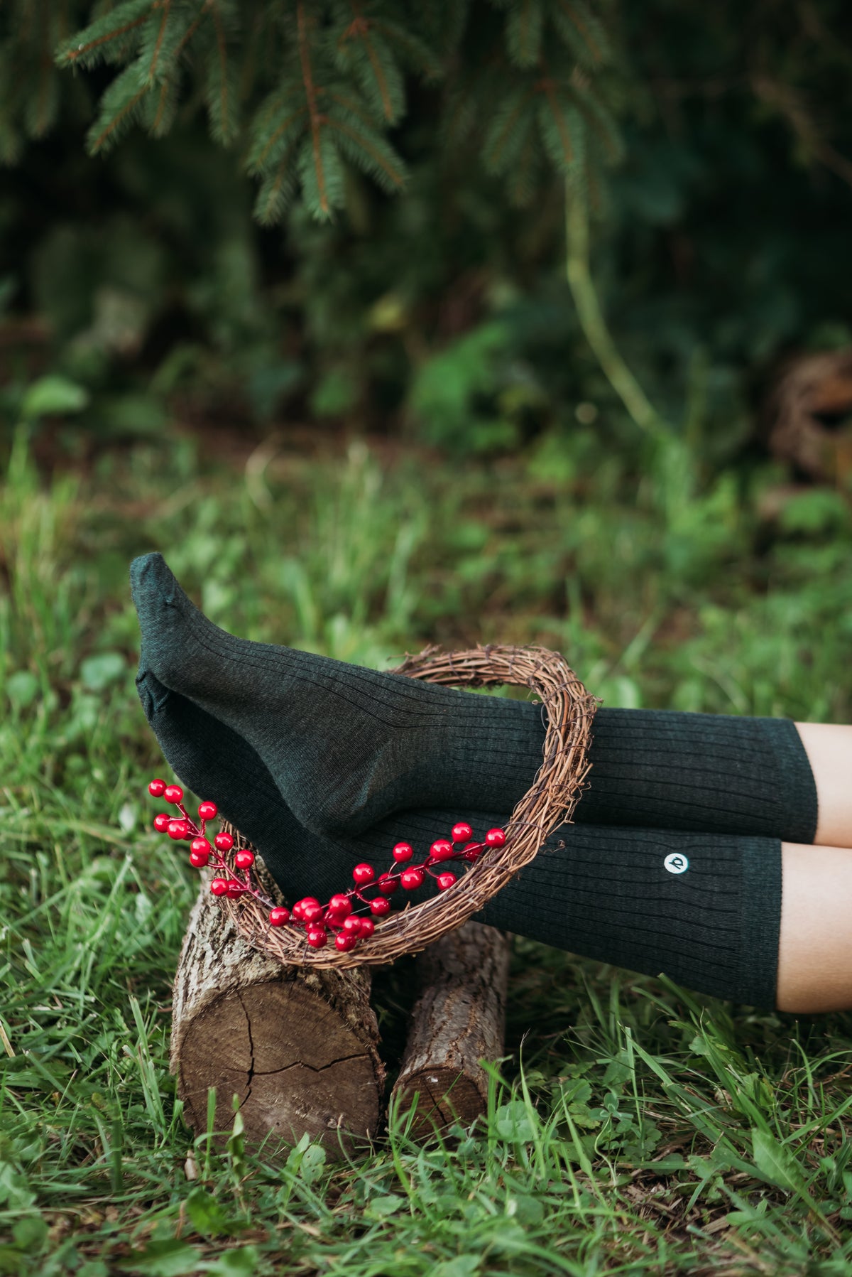 Black ribbed crew socks worn on a person's legs, resting on a log outdoors on green grass with a grapevine wreath and red berries in the foreground.