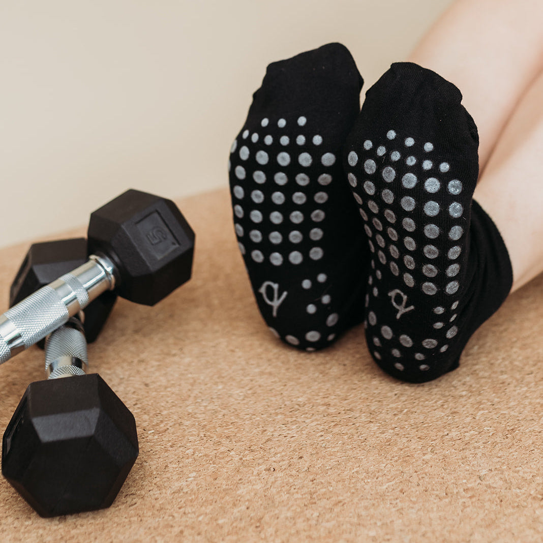 Black grip socks with silver non-slip dots worn on a person's feet, shown with dumbbells on a cork mat against a beige background.