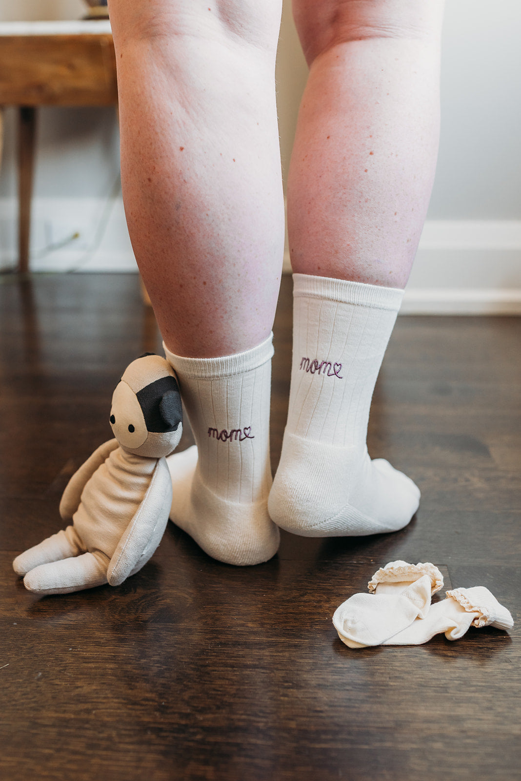 Person wearing white socks with text, standing on a wooden floor next to a plush turtle toy.