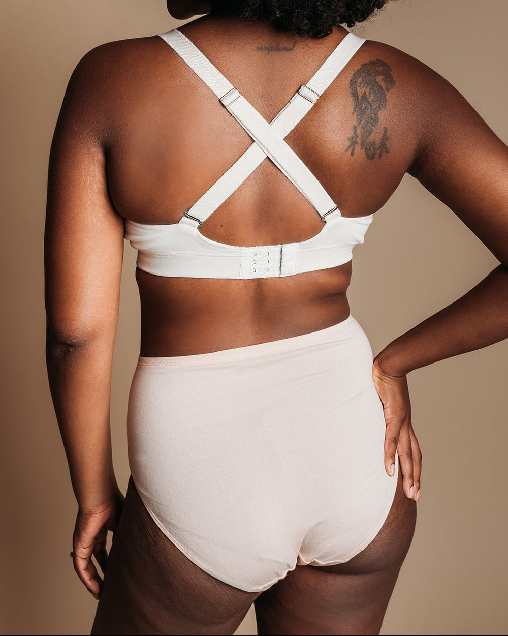 A woman shown from the back wearing a white high-waisted bra and matching high-waisted briefs against a warm beige studio background.