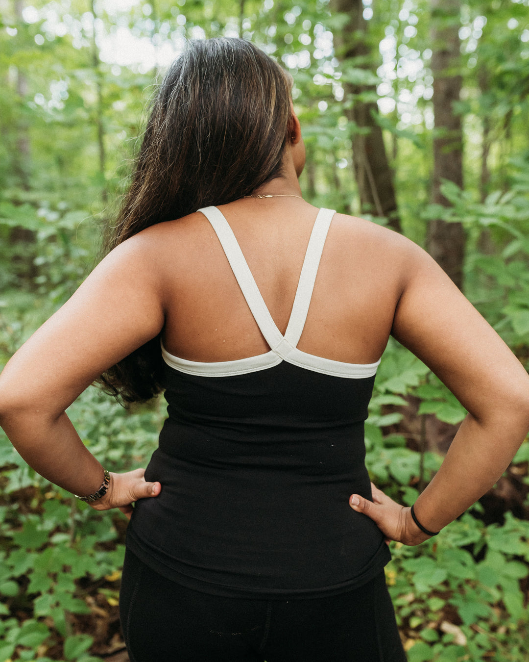 A woman viewed from behind wearing a black athletic tank top with contrasting white crossover shoulder straps and a scoop neckline, set against a lush green forest background.