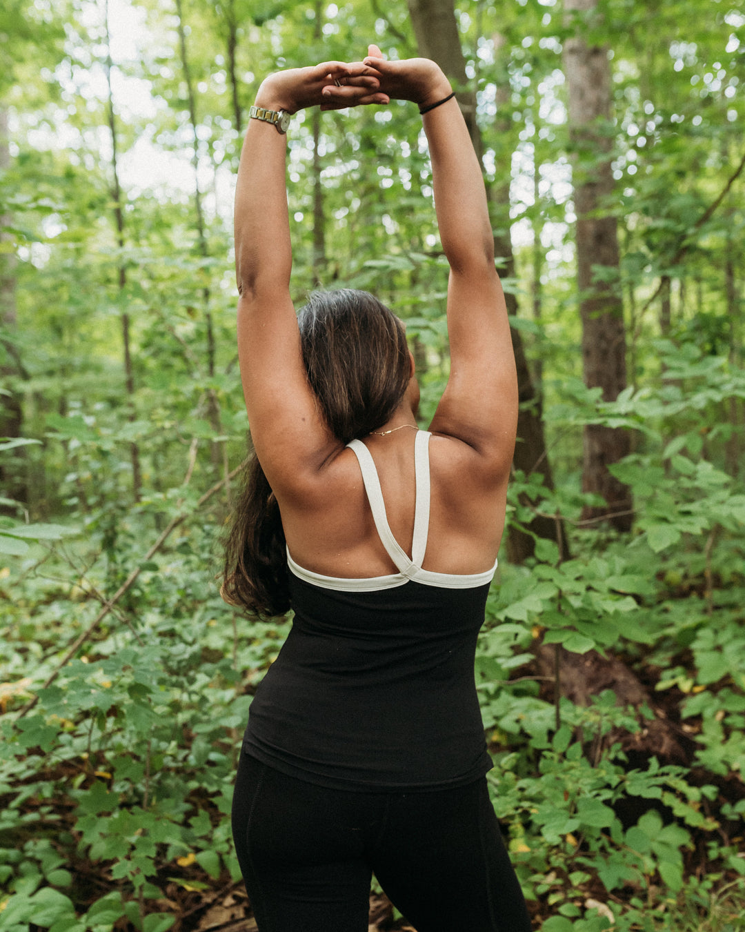 A woman viewed from behind in a forest, wearing a black athletic tank top with white crossover shoulder straps and black leggings, stretching her arms overhead.