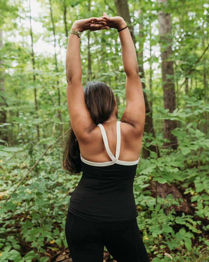 A woman viewed from behind in a forest, wearing a black athletic tank top with white crossover shoulder straps and black leggings, stretching her arms overhead.