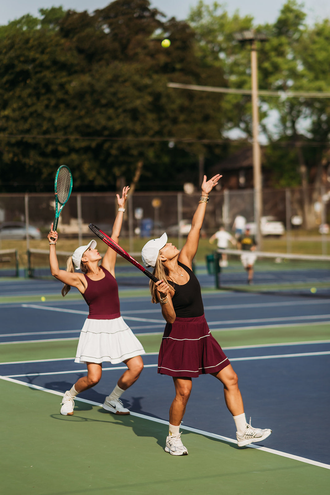 Two women playing tennis on an outdoor hard court, one serving and reaching high while the other prepares with a racket. Both wear matching dark maroon athletic tops and skirts, with one in white and the other in black, against a blue court and green trees in the background.