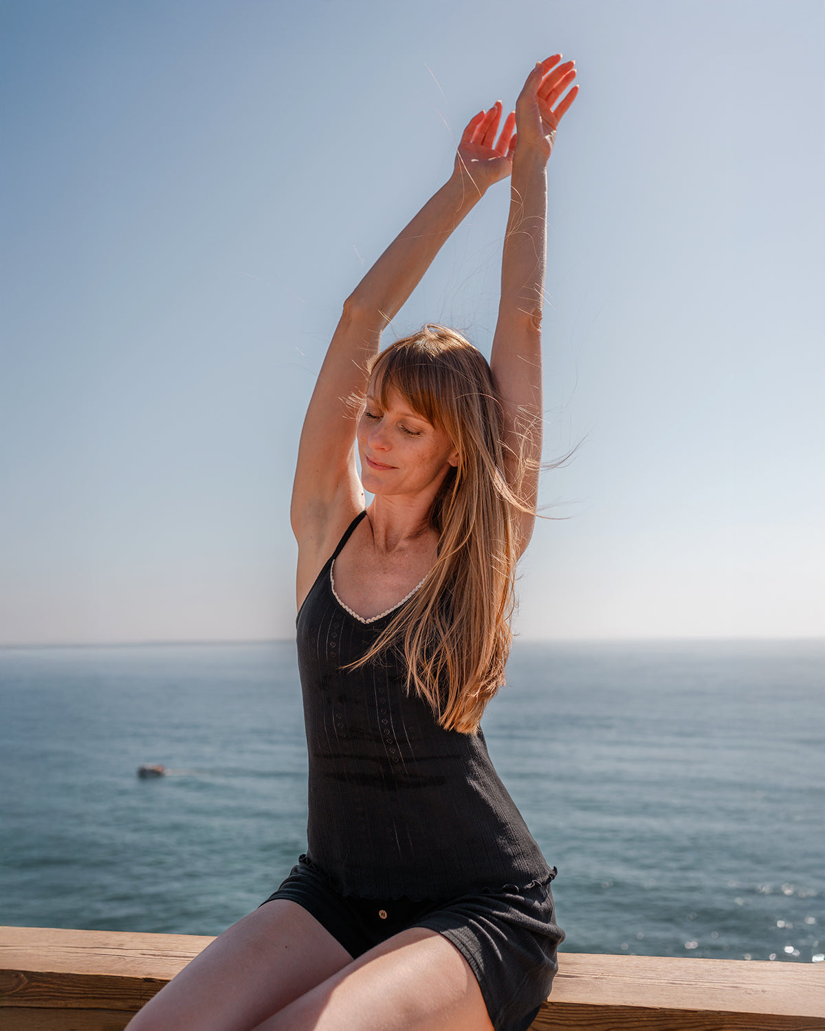 Woman sitting oceanside with arms raised in Women's Pointelle Organic Cotton Tank Top in Black | Q for Quinn